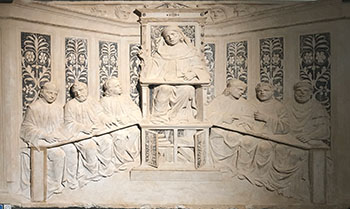 The image shows a carving of medieval scene in stone, that depicts the wise lecturer departing knowledge on to his students. Taken at Bologna Medieval museum.